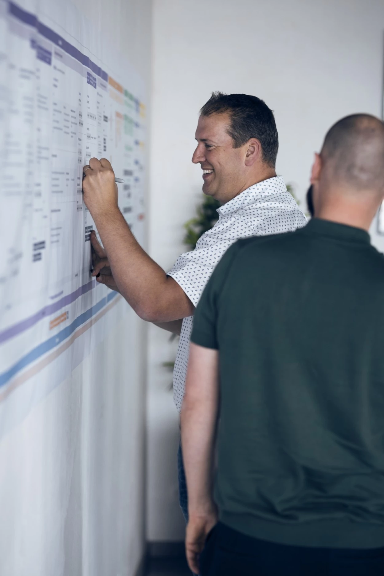 Two professionals evaluating a project timeline on a wall-mounted board in an office setting.
